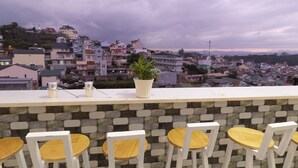 A bar with white chairs and a potted plant, overlooking a cityscape with various buildings and a cloudy sky. - MrLee Dalathouse (Xuan Huong - Da Lat)