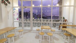 A dining area with multiple wooden tables and white stools, large windows with a view of buildings and a cloudy sky. - MrLee Dalathouse (Xuan Huong - Da Lat)