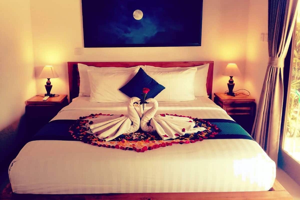 A hotel room with a bed, two bedside tables, a nightstand, a painting of a moon, and a window with curtains.