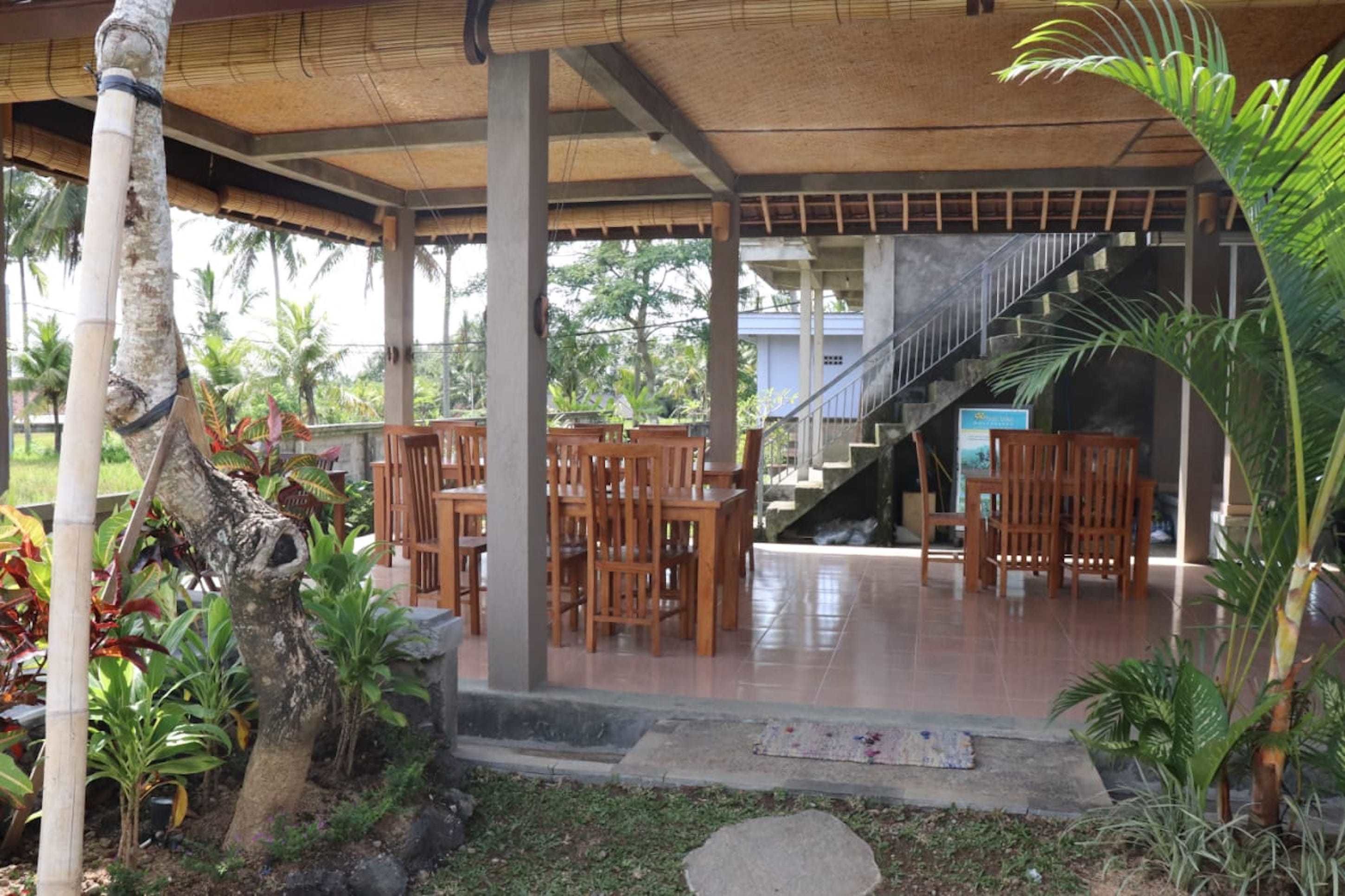 An outdoor covered seating area with wooden chairs and tables, surrounded by tropical plants and a staircase leading to an upper level.