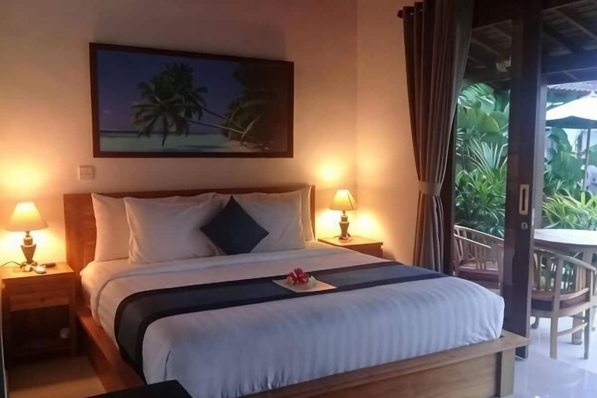 A hotel room with a bed, two bedside lamps, a nightstand, a painting of a beach scene, and a view of the outdoors.