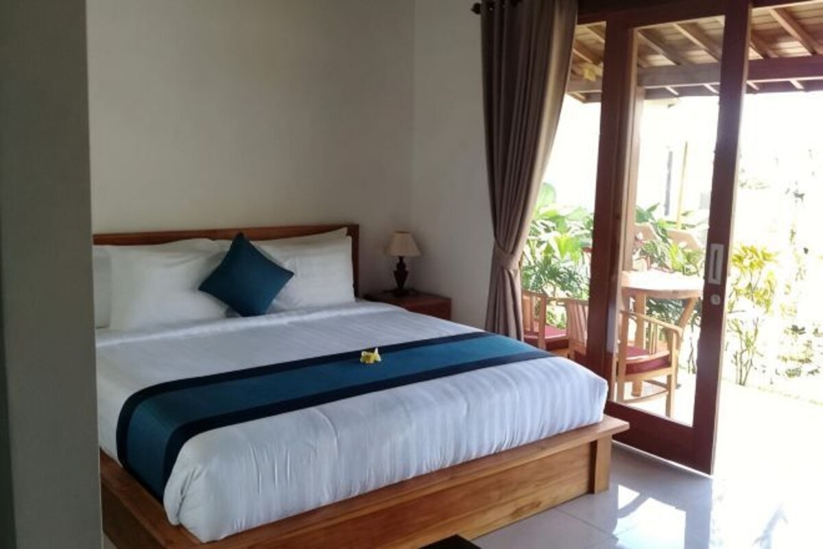 Una cama individual con estructura de madera, una colcha blanca y azul, una mesita de noche con lámpara, y vistas al exterior a través de una puerta de vidrio.