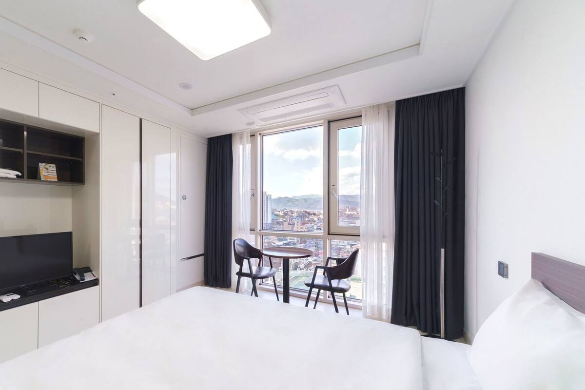 A hotel room with a large bed, a desk with two chairs, a TV, and a window with a city view.