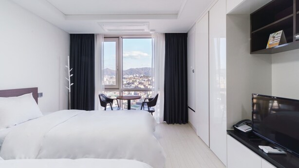 A hotel room with two beds, a desk with a phone and a TV, a window with a city view, and a small table with chairs. - The Venezia Suite Hotel (Chuncheon)
