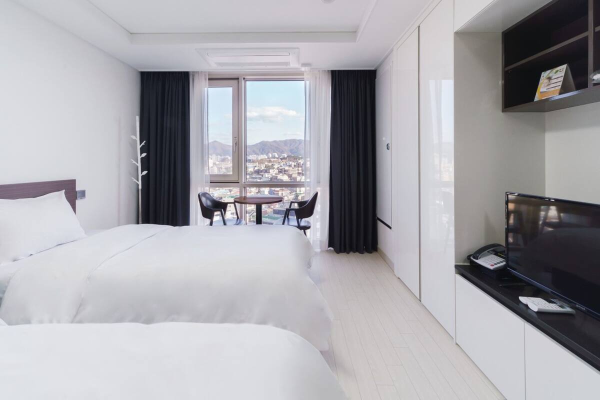 A hotel room with two beds, a desk with a phone and a TV, a window with a city view, and a small table with chairs.
