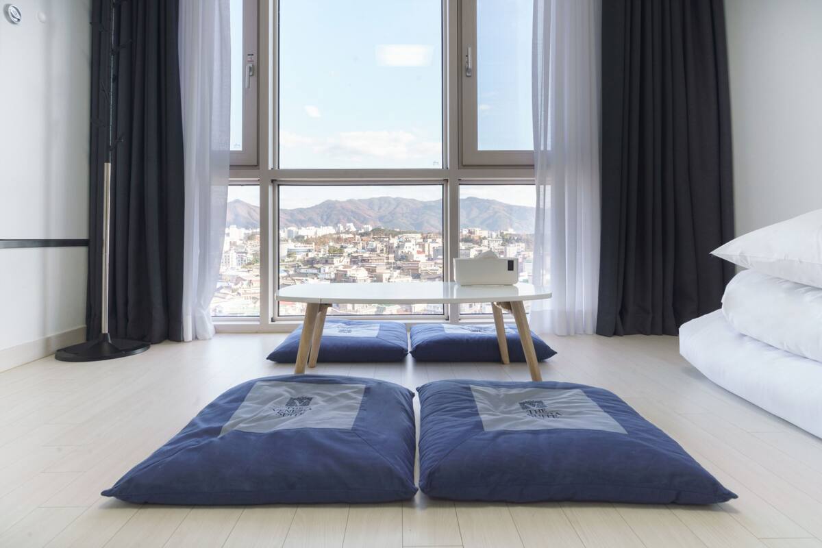 A modern hotel room with a large window offering a city view, a white bed, and a small table with blue cushions.