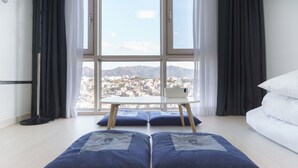 A modern hotel room with a large window offering a city view, a white bed, and a small table with blue cushions. - The Venezia Suite Hotel (Chuncheon)