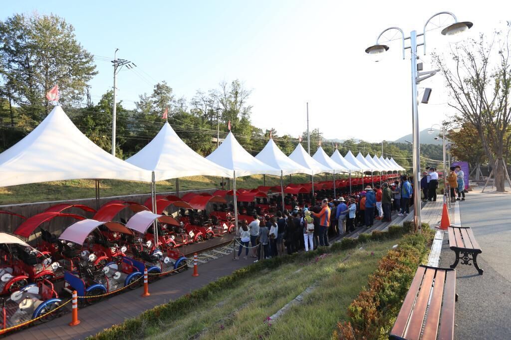 A row of white tents with red canopies, a group of people gathered, and red vehicles parked in front.