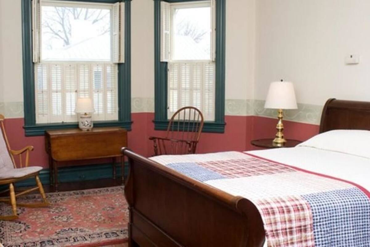 A bedroom with a wooden bed, two rocking chairs, a desk with a lamp, and a window with shutters.