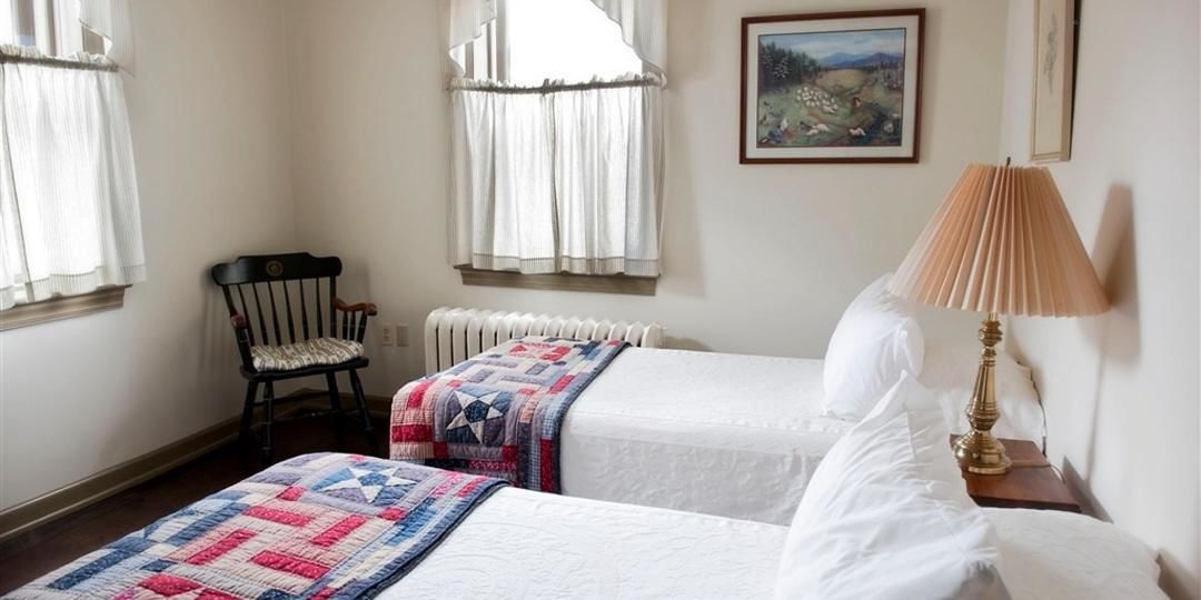 A bedroom with two beds, a chair, a radiator, a lamp, and a painting on the wall.