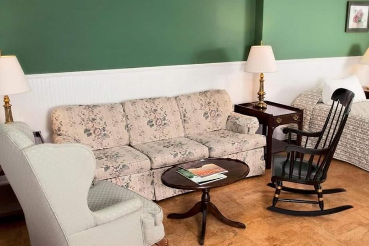 A room with a green wall, a floral sofa, a rocking chair, a small table with a book, and two bedside tables with lamps.