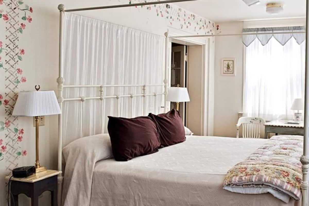 A four-poster bed with a canopy, a bedside table with a lamp, a chair, and a desk with a chair in a bedroom.