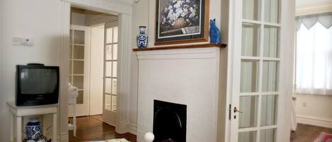Una habitación con chimenea, un televisor sobre un mueble bajo, una puerta que da a otra habitación y un cuadro enmarcado sobre la chimenea.