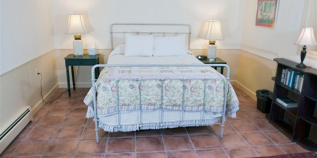 A neatly arranged bedroom with a bed, two bedside tables with lamps, a small bedside table with a lamp, a bookshelf, and a radiator.