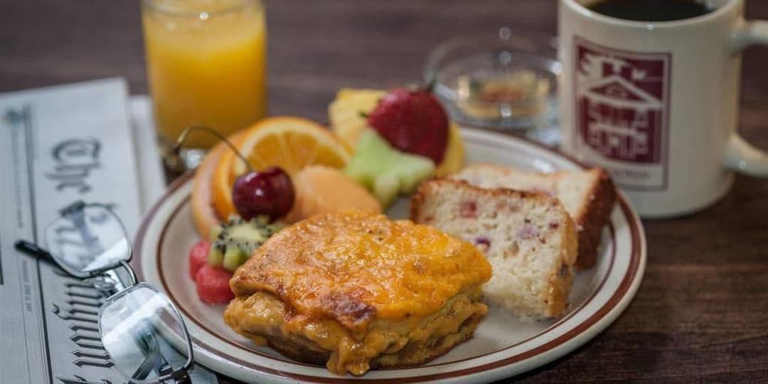 Un plato con un sándwich de desayuno, tostadas y fruta, un vaso de jugo de naranja y una taza de café sobre una mesa de madera.