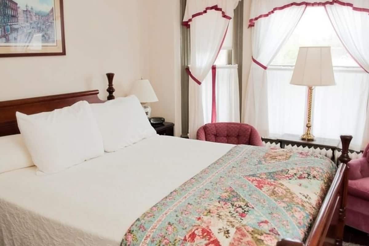 A neatly made bed with white linens and a floral bedspread, a wooden headboard, a nightstand with a lamp, and a pink armchair.
