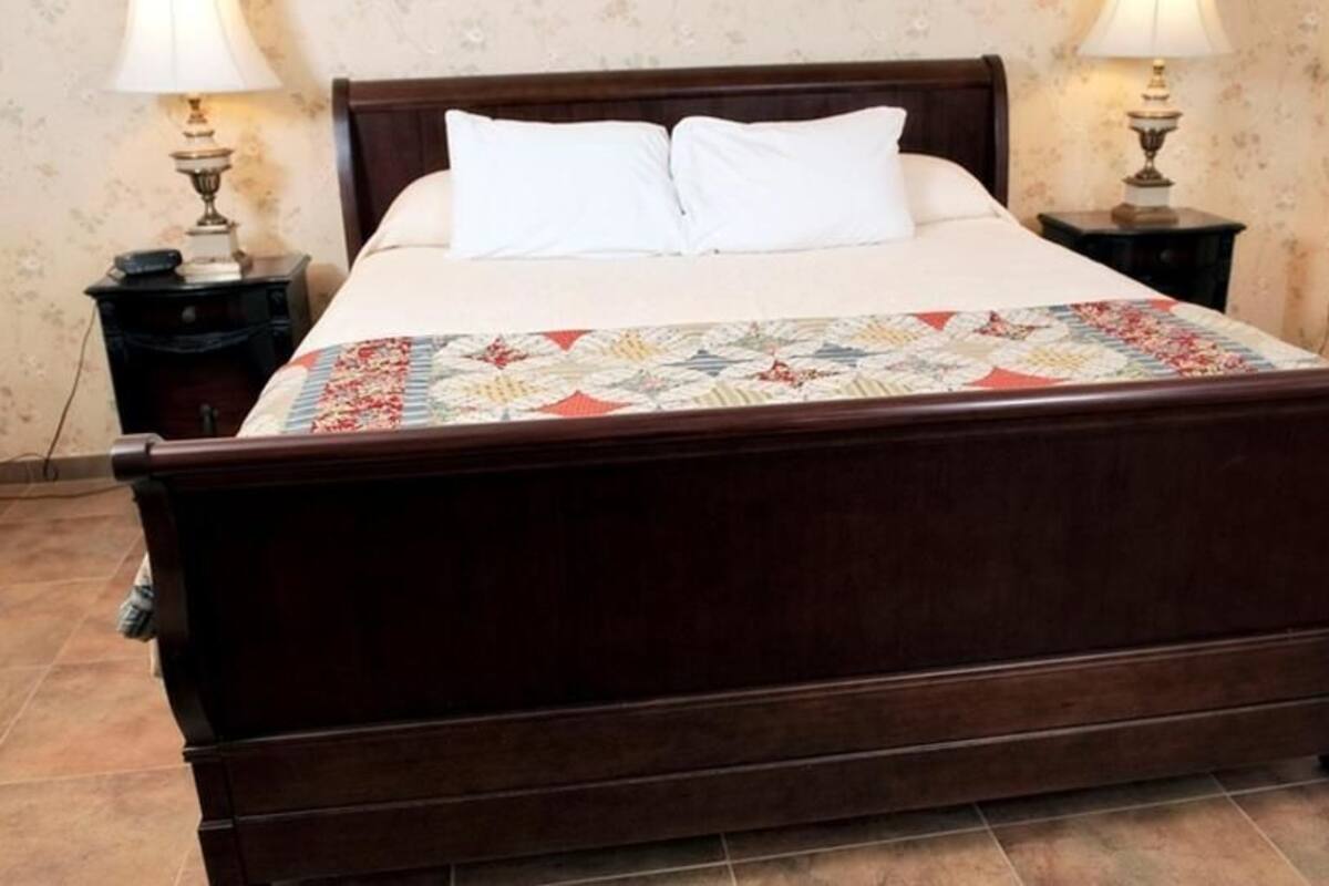 A wooden bed with white pillows and a colorful quilt, flanked by two bedside tables with lamps, in a room with floral wallpaper.