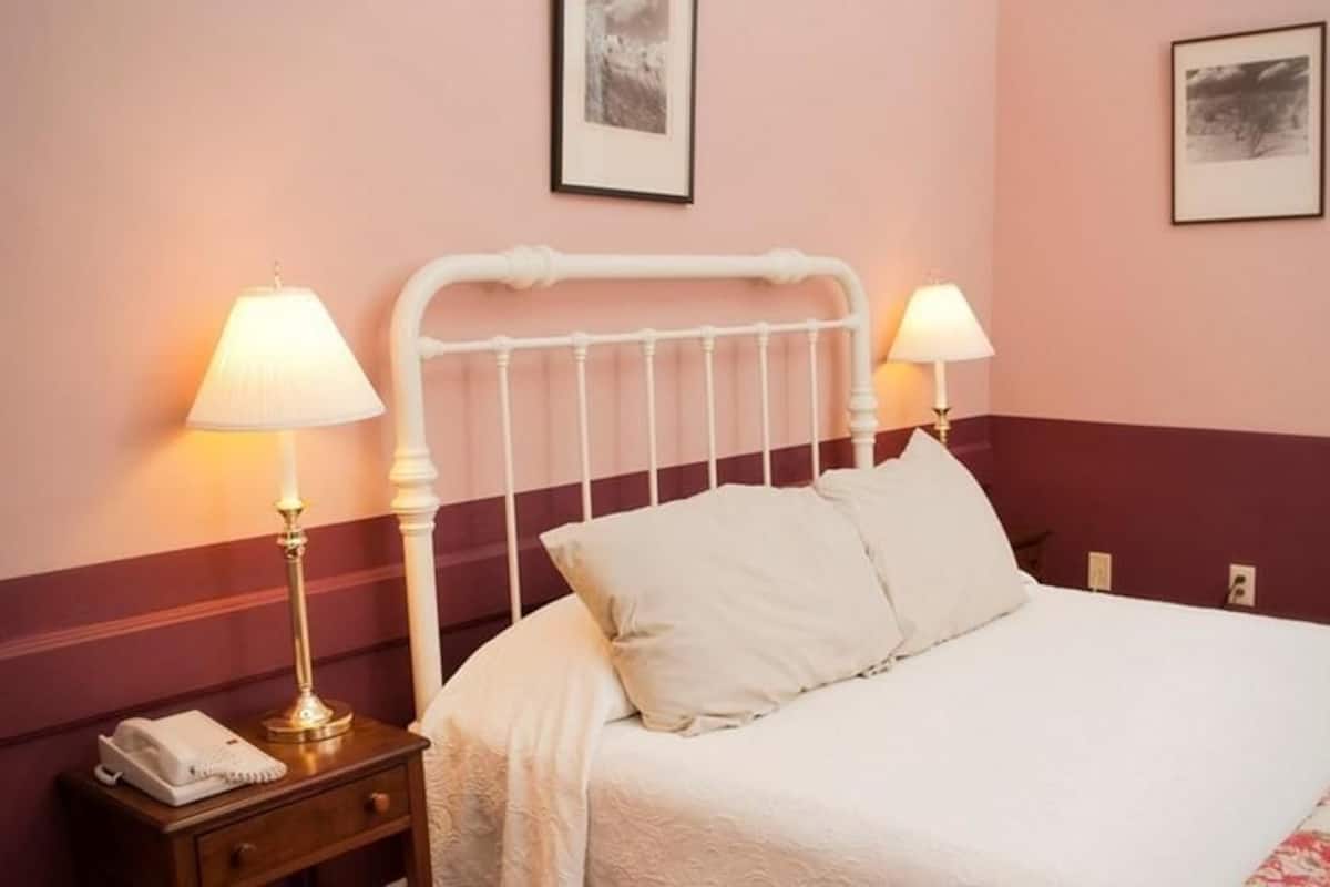 A neatly made bed with white linens and two pillows, a bedside table with a lamp and a telephone, and two framed pictures on the wall.