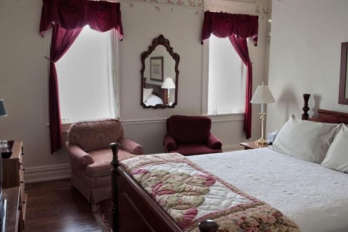 A hotel room with a large bed, two armchairs, a wooden nightstand, a mirror, and a framed picture on the wall.