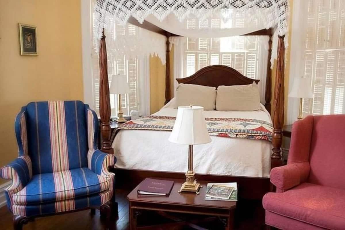 A four-poster bed with a canopy, a striped armchair, a red armchair, and a bedside table with a lamp and books.