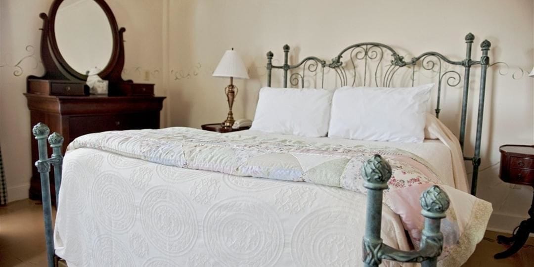Una cama bien hecha con una colcha blanca y estampada, un aparador de madera con espejo y una mesita de noche con lámpara.