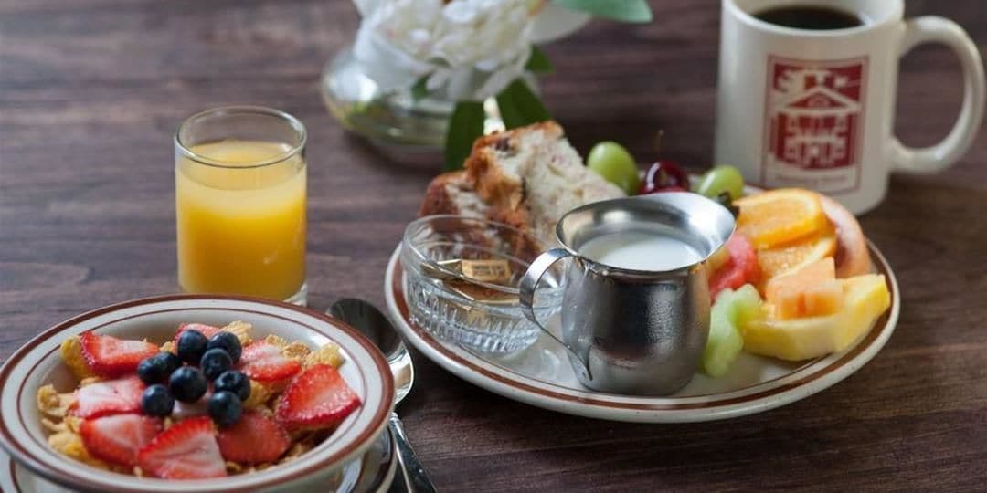 A breakfast setting with a bowl of cereal topped with strawberries and blueberries, a glass of orange juice, a plate of fruit, a cup of coffee, and a small metal cup.