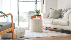 A modern living room with a white sofa, a round ottoman, a wooden side table with a plant, and a view through a glass door. - Cosy 2 Bedroom Villa with super comfortable KING BEDS & luxury linen. Near Beach (Coffs Harbour)