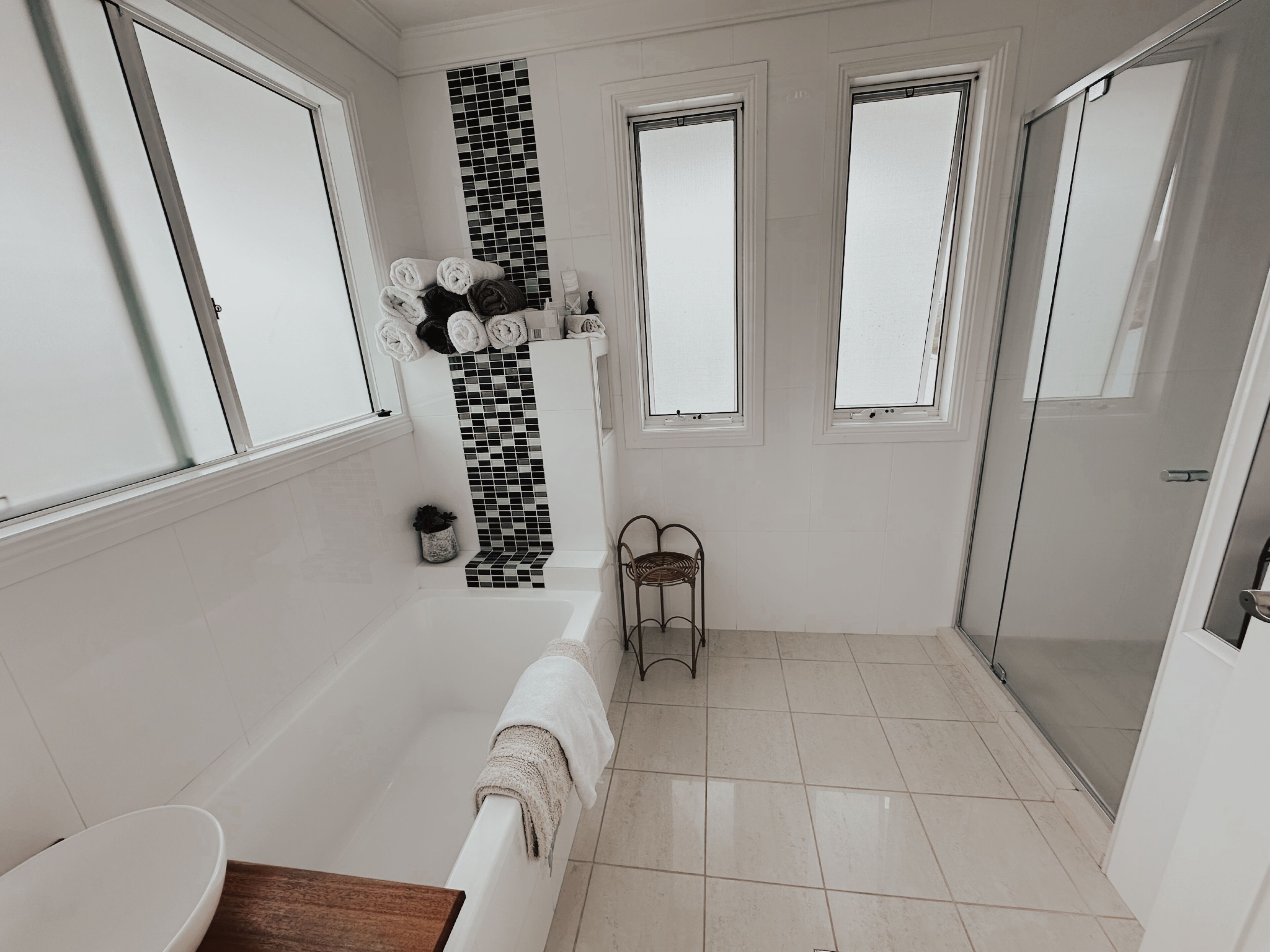 Une salle de bain moderne avec une baignoire, une douche et un lavabo. On y trouve des serviettes pliées et posées sur un étagère murale, une petite plante en pot et une chaise.