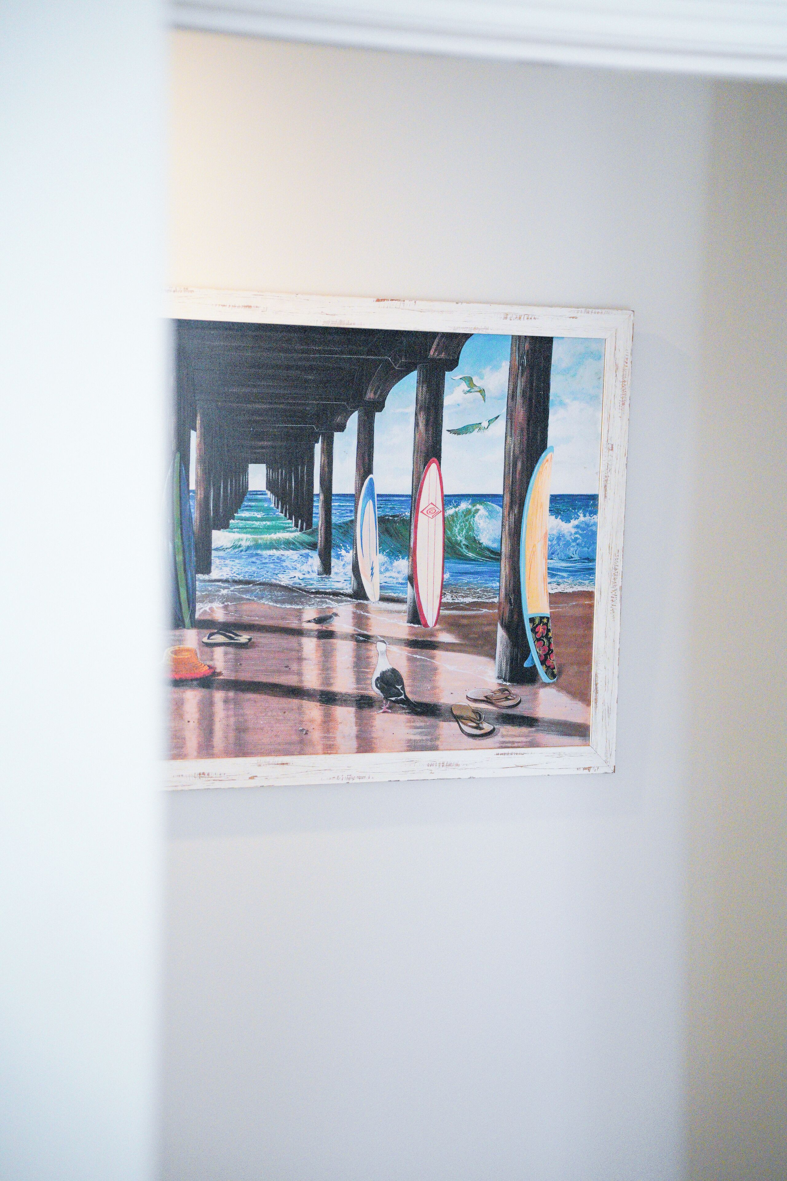 A framed artwork of a pier with surfboards and a bird, mounted on a wall.