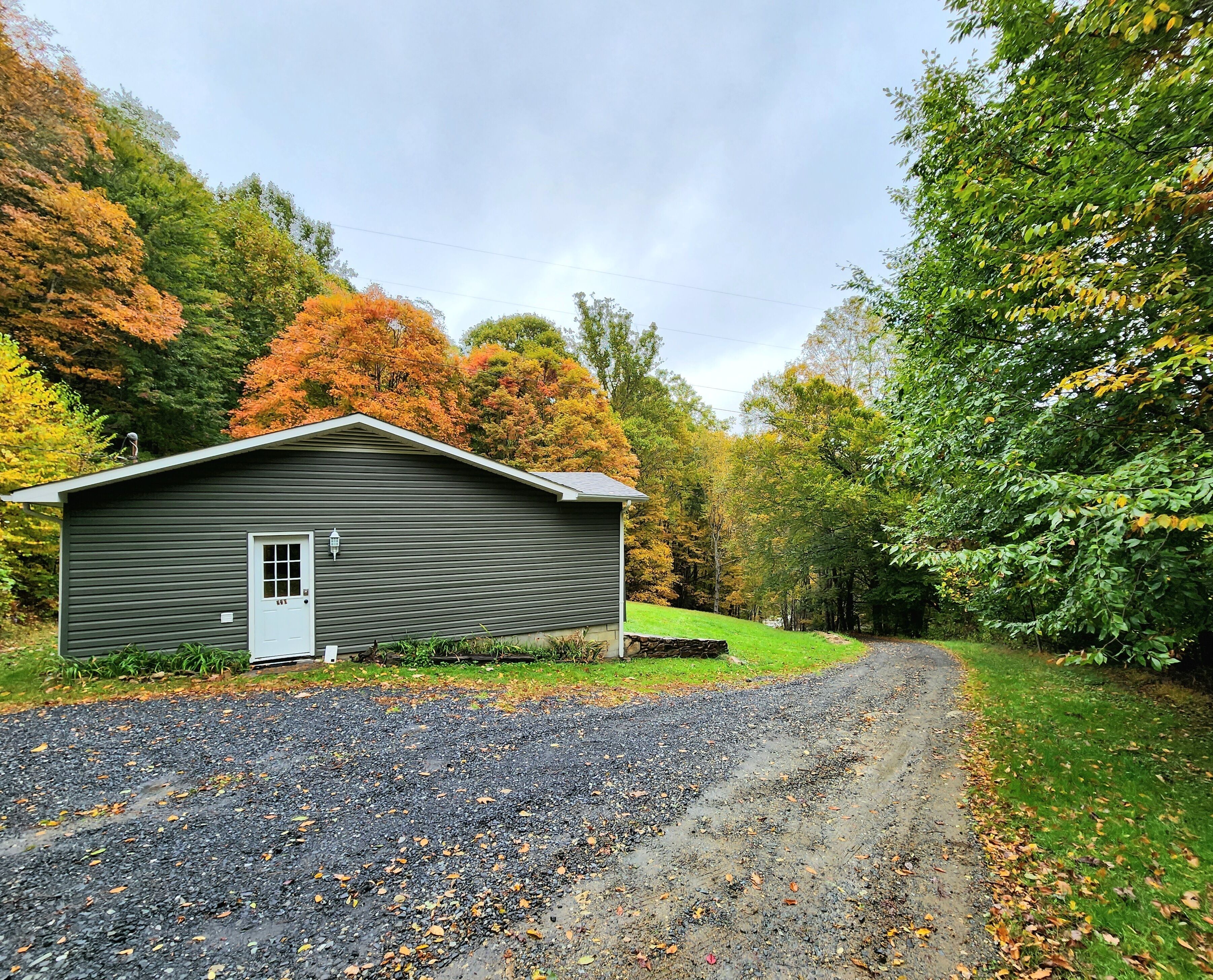 A small, single-story cabin with a white door and window, surrounded by a gravel driveway and lush autumn foliage.
