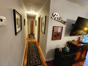 A hallway with framed pictures, a wooden floor, a rug, a television, and a cabinet with a kettle.
