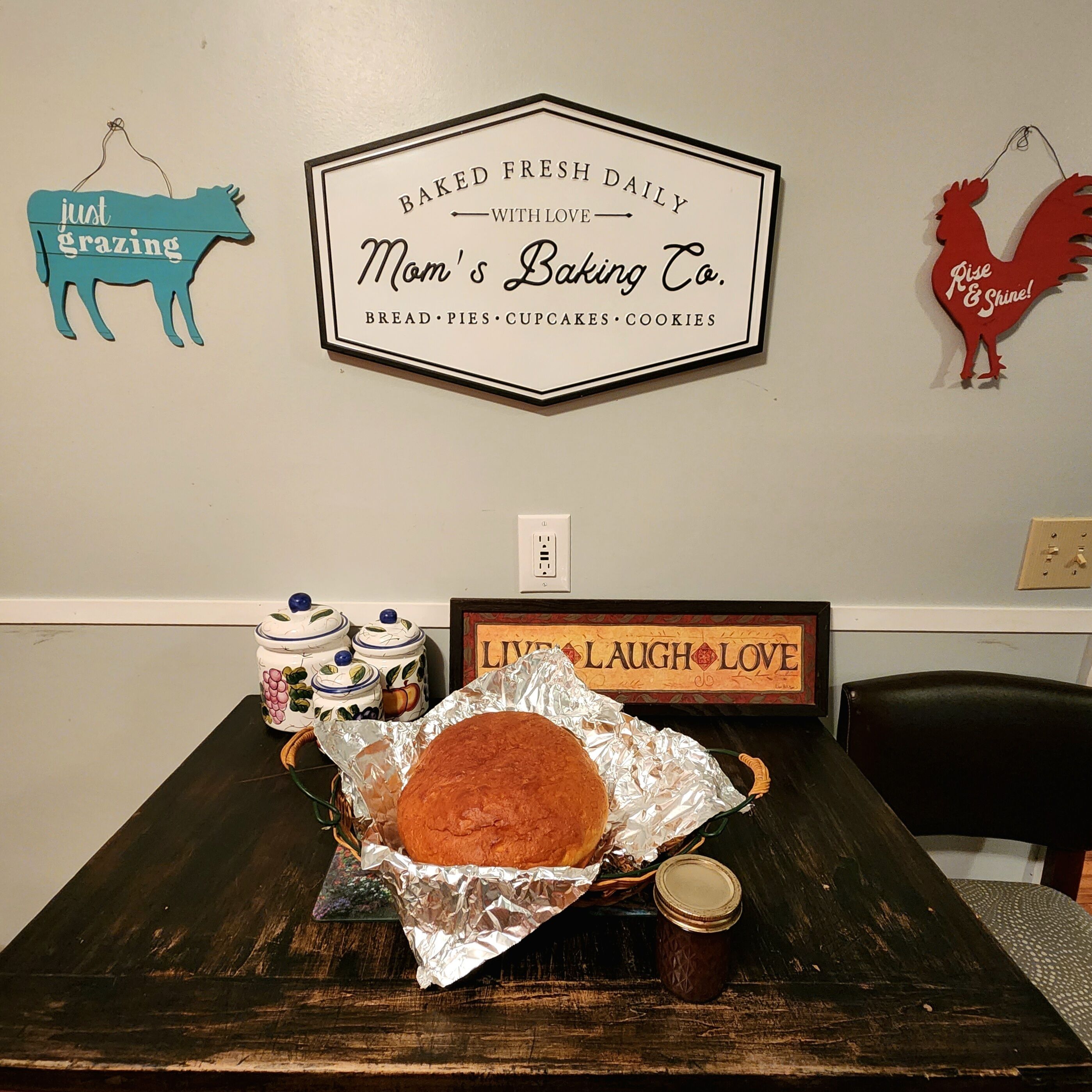 A kitchen with a loaf of bread on a table, surrounded by jars and a sign that reads "Baked Fresh Daily with Love Mom's Baking Co."