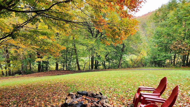 Un espace extérieur aménagé avec des sièges, un foyer et des chaises rouges, le tout entouré d’arbres aux feuilles d’automne.