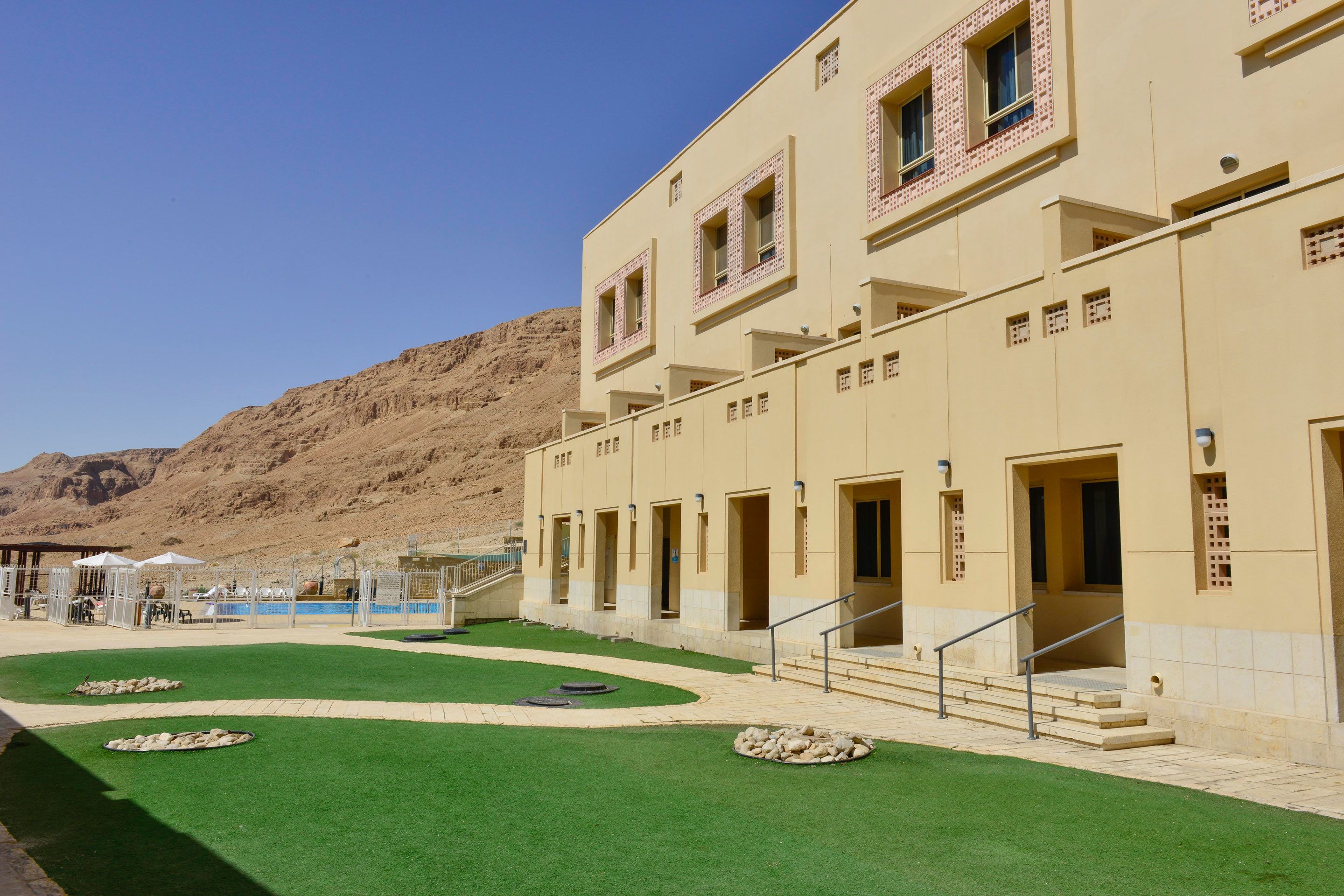 Un hotel con techo plano, varios balcones y un patio ajardinado con césped artificial y piscina.