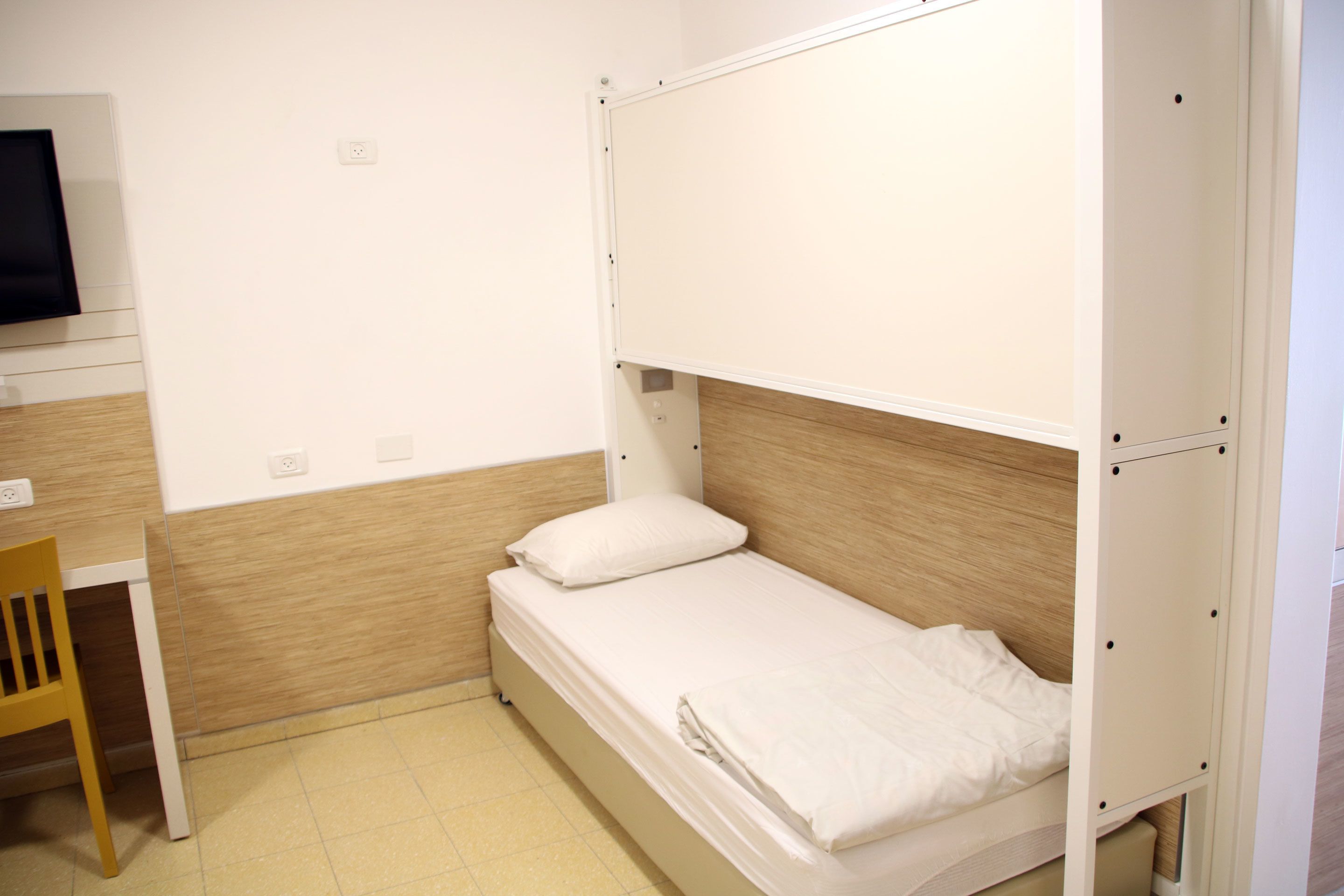 Una habitación pequeña con una cama individual, cabecera de madera, un escritorio con silla y televisión en la pared.