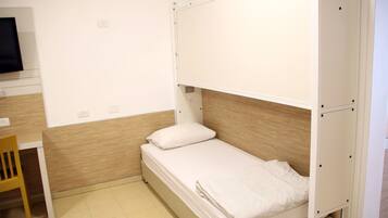 Una habitación pequeña con una cama individual, cabecera de madera, un escritorio con silla y televisión en la pared.