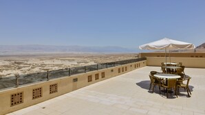 A terrace with a table and chairs, a white umbrella, and a view of a desert landscape. - HI Masada Hostel (Tamar)
