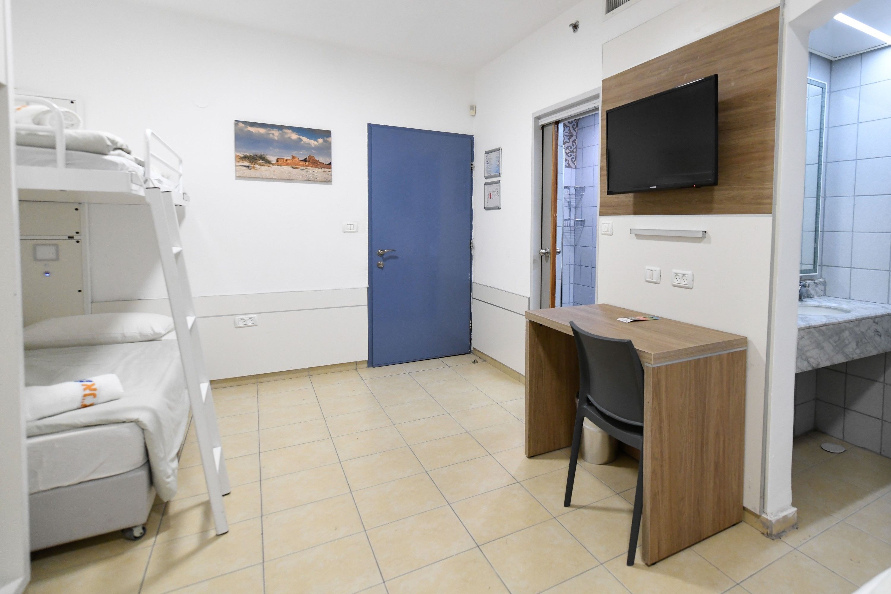 A room with a bunk bed, a desk with a chair, a television mounted on the wall, and a bathroom with a marble sink.