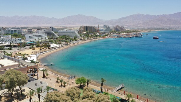 A coastal cityscape with a beach, buildings, and mountains in the background. - HI Eilat Hostel (Eilat)