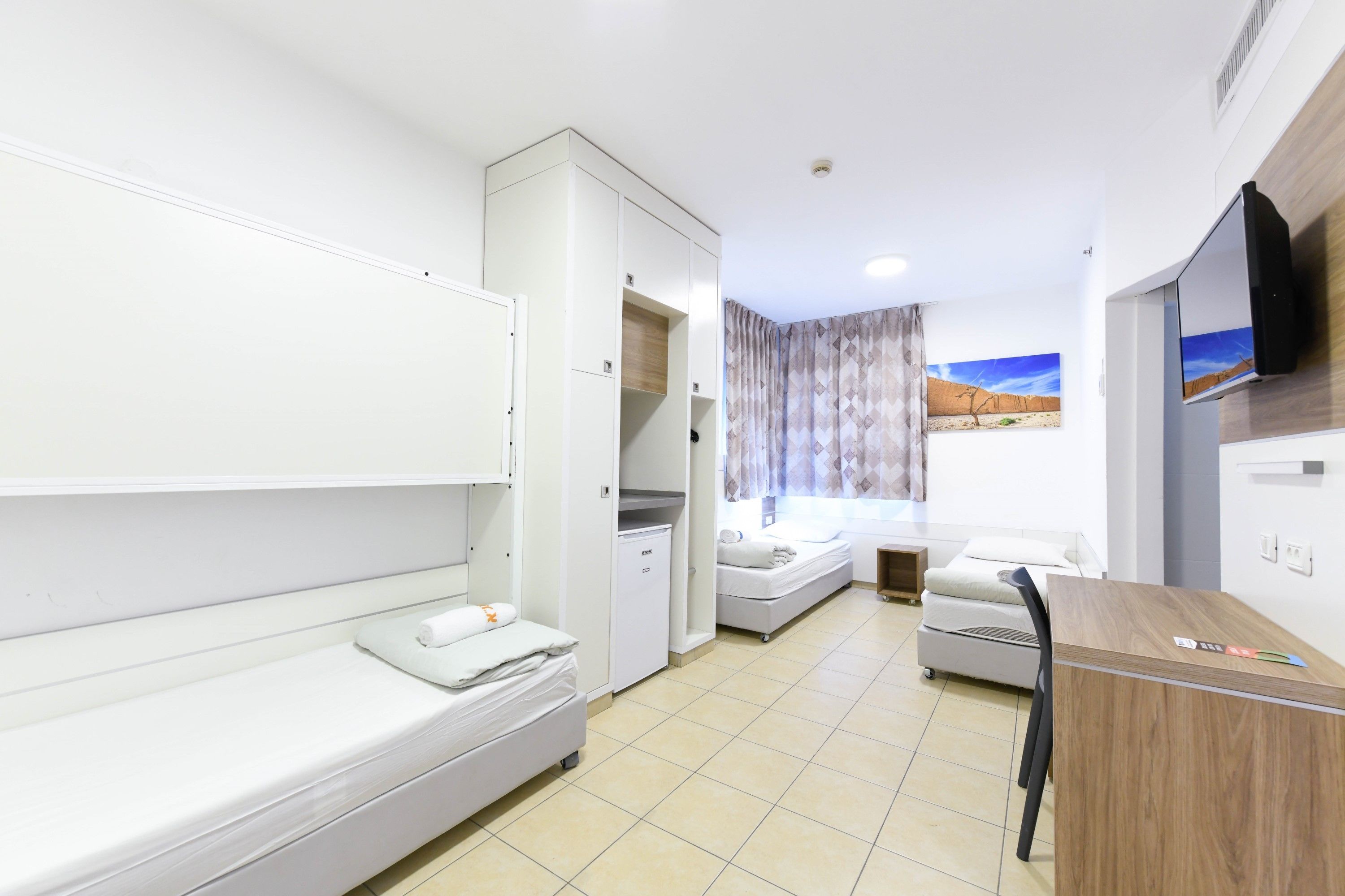 Een kleine hotelkamer met een enkel bed, een bureau, een kledingkast en een aan de muur bevestigde televisie.