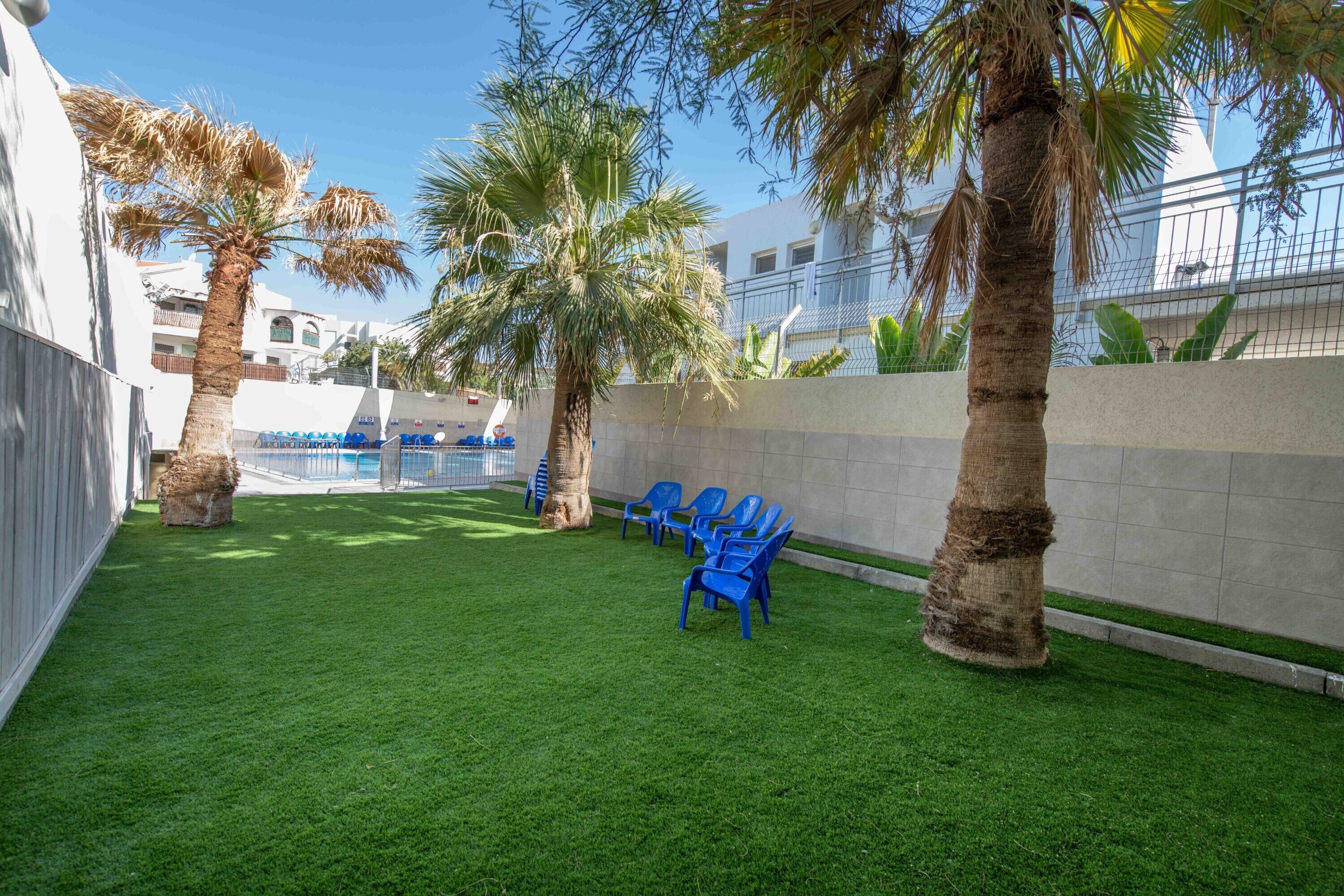 A courtyard with artificial grass, palm trees, and blue plastic chairs.