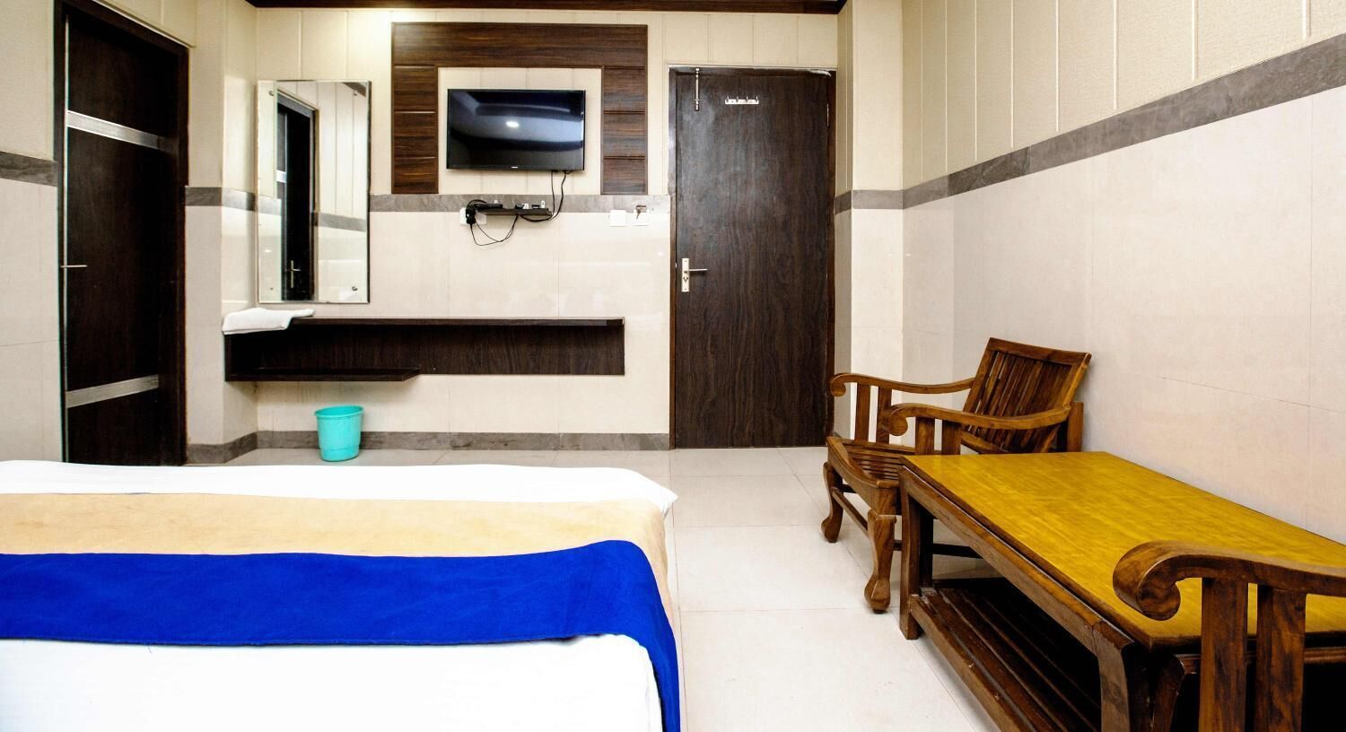 A hotel room with a bed, a wooden table, two chairs, a television mounted on the wall, and a door.
