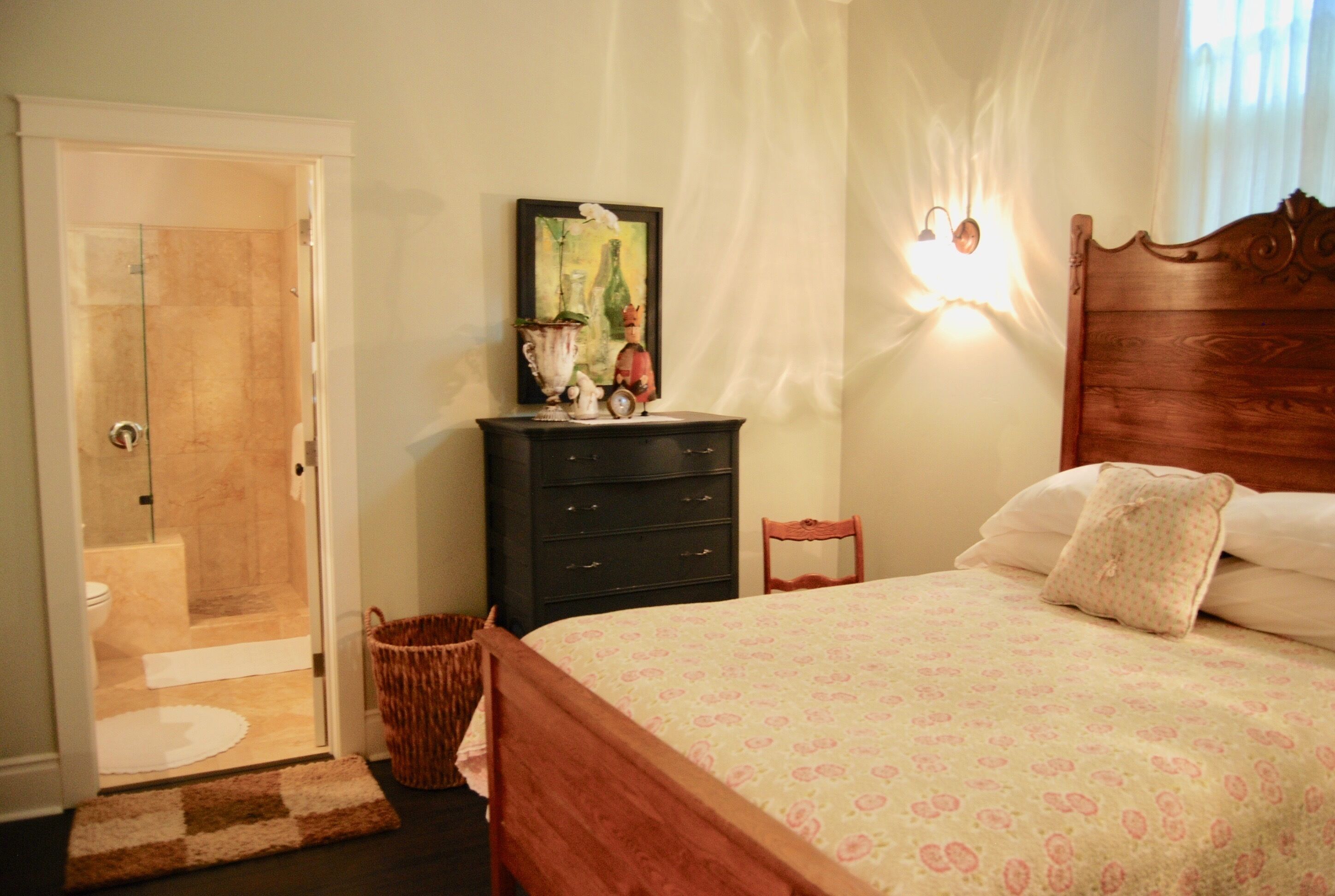 Un dormitorio con cama, cómoda, silla, lámpara de pared y puerta que da al baño.