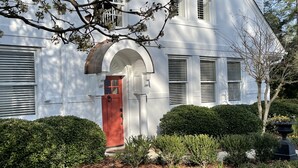 Una casa blanca con puerta roja, contraventanas blancas y un jardín cuidado.
