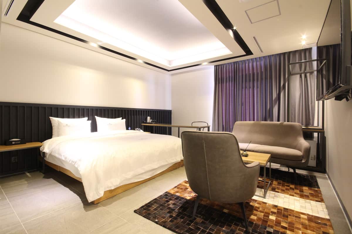 A modern hotel room with a large bed, a seating area with a sofa and armchair, a small table, and a patterned rug.