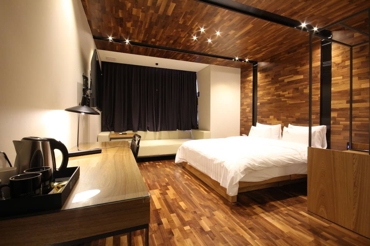 A modern hotel room with a wooden ceiling, a bed, a sofa, a desk with a coffee maker, and a mirror.