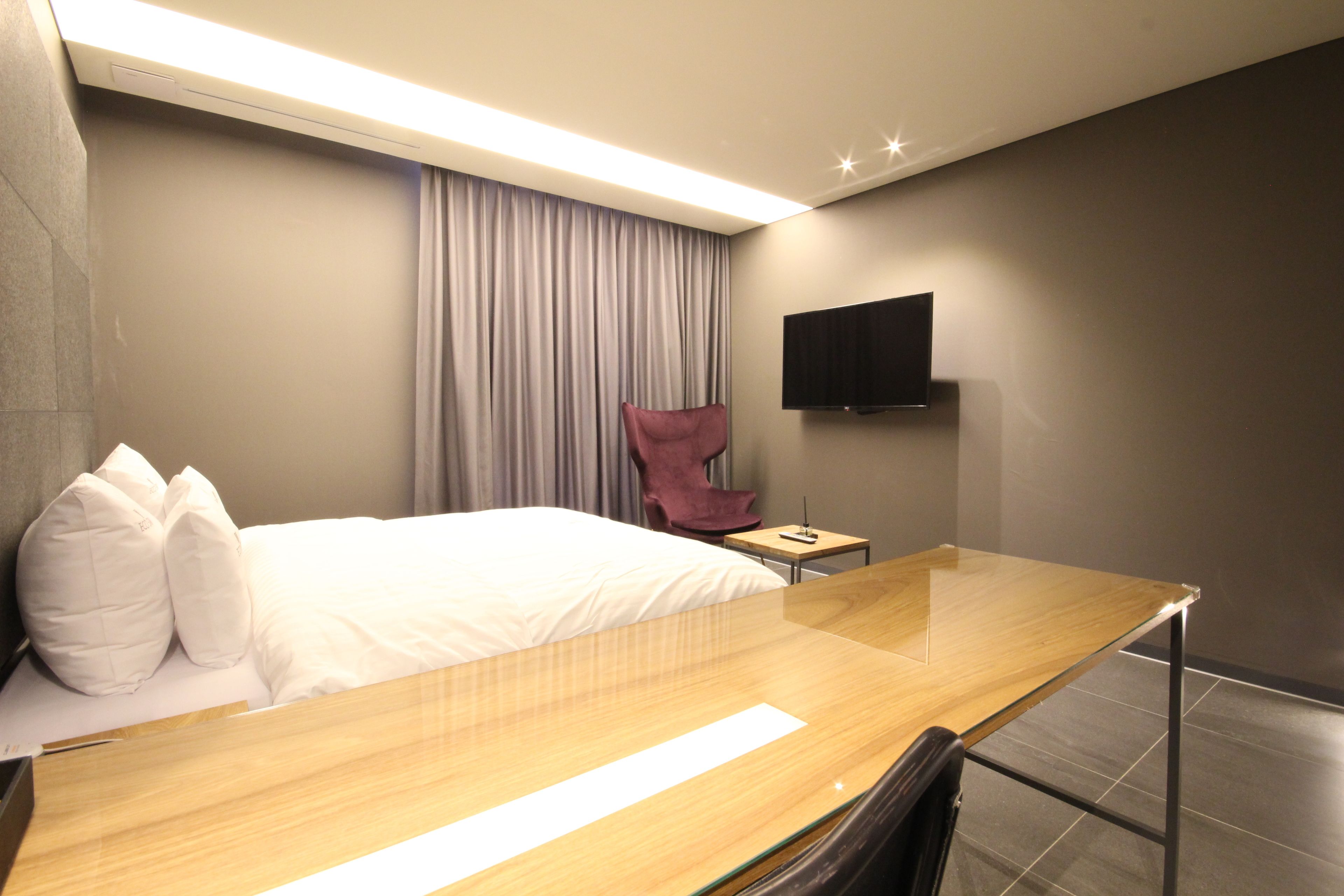 A modern hotel room with a wooden bed, a flat-screen TV mounted on the wall, a small table, and a chair.