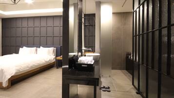 Una habitación de hotel moderna con una cama grande, un baño con lavamanos y espejo, y un tabique de vidrio.