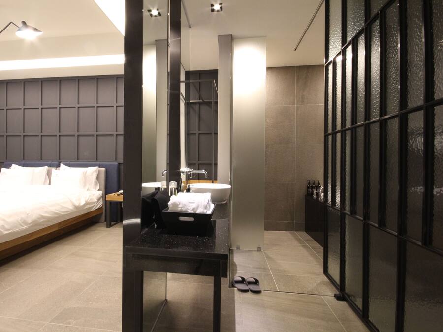 Una habitación de hotel moderna con una cama grande, un baño con lavamanos y espejo, y un tabique de vidrio.