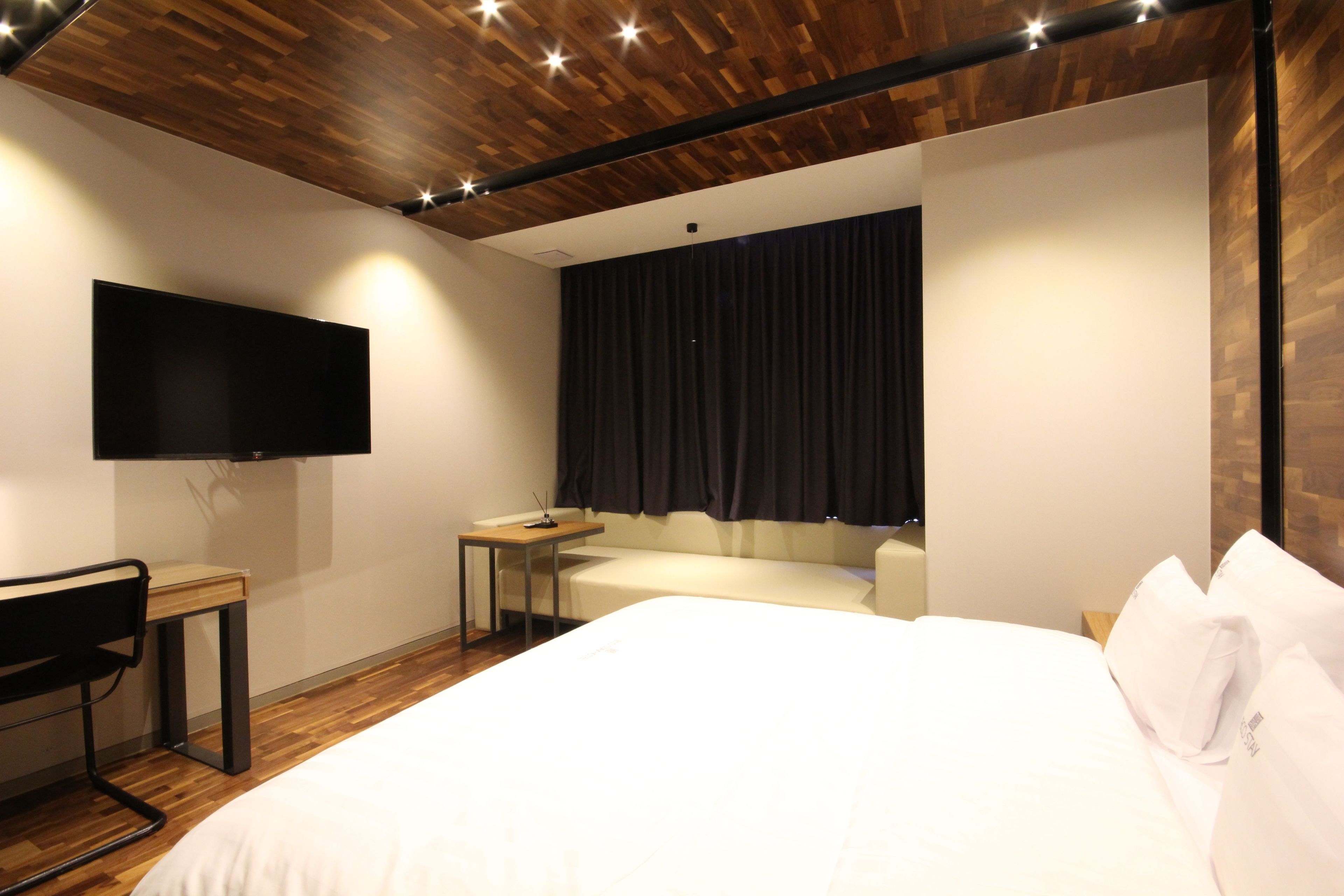 A modern hotel room with a wooden ceiling, a flat-screen TV mounted on the wall, a bed with white bedding, a small desk, and a chair.